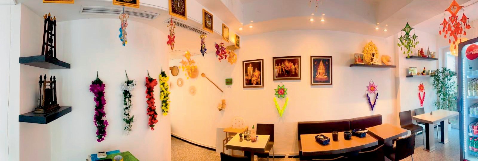 Das Lokal innen: weiße Wände mit Blumenketten, gerahmten Bildern und Schmuck aus Thailand, Tische und eine Sitzbank